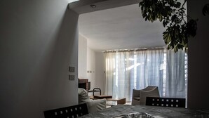 Un coin repas avec une table dressée pour deux personnes, une chaise et une table à manger recouverte d’une nappe à motifs. Une fenêtre avec des rideaux légers laisse entrer la lumière, et une plante est suspendue au-dessus de la table.