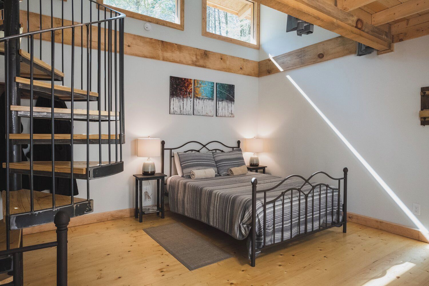 Un dormitorio con una cama, dos lámparas de noche, suelo de madera, una escalera espiral de metal negro y un cuadro en la pared.