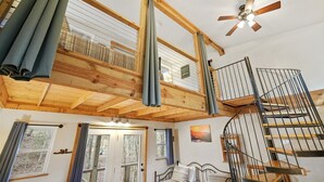 A wooden loft bedroom with a spiral staircase, a ceiling fan, and large windows. - Fairytale SQUIRREL'S NEST cabin at Mulberry Gap in Ellijay! (Ellijay)