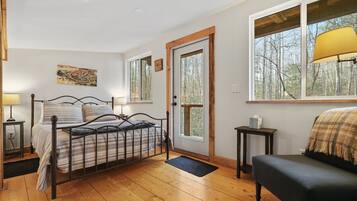 Une chambre avec un lit en métal, un plancher en bois, une porte avec une fenêtre, une table de chevet avec une lampe, une chaise et une vue sur les arbres par la fenêtre.