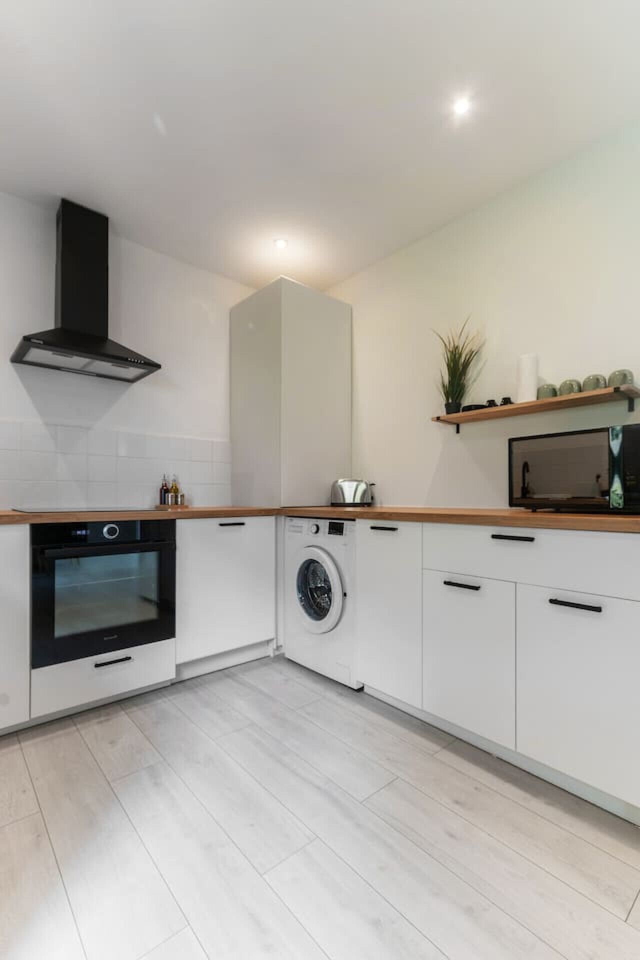 Een moderne keuken met witte kastjes, een inbouwoven, een wasmachine en een magnetron op een houten aanrecht.