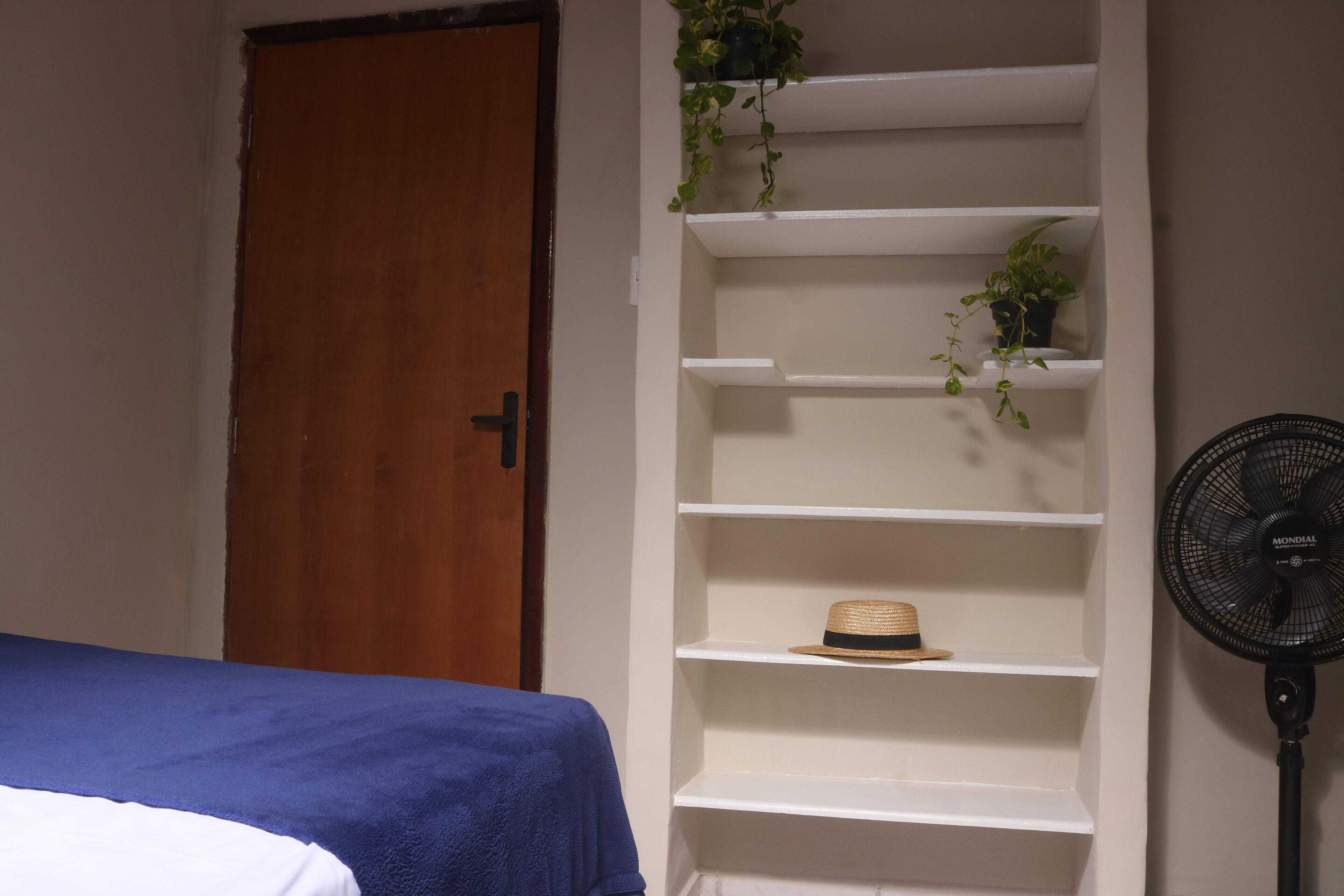 Una camera da letto con un letto, un mobiletto contenitore bianco con piante, un cappello e una ventola da pavimento.