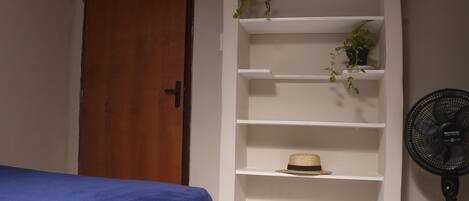 Una camera da letto con un letto, un mobiletto contenitore bianco con piante, un cappello e una ventola da pavimento.