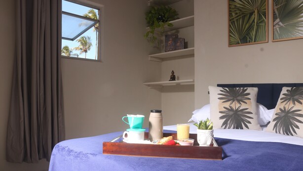Una cama bien hecha con una colcha azul, una bandeja con alimentos para el desayuno, una ventana con cortinas y una estantería con objetos decorativos.