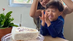 Una mujer y un niño en una mesa con un pastel, una planta en maceta y una ventana con vista a follaje.