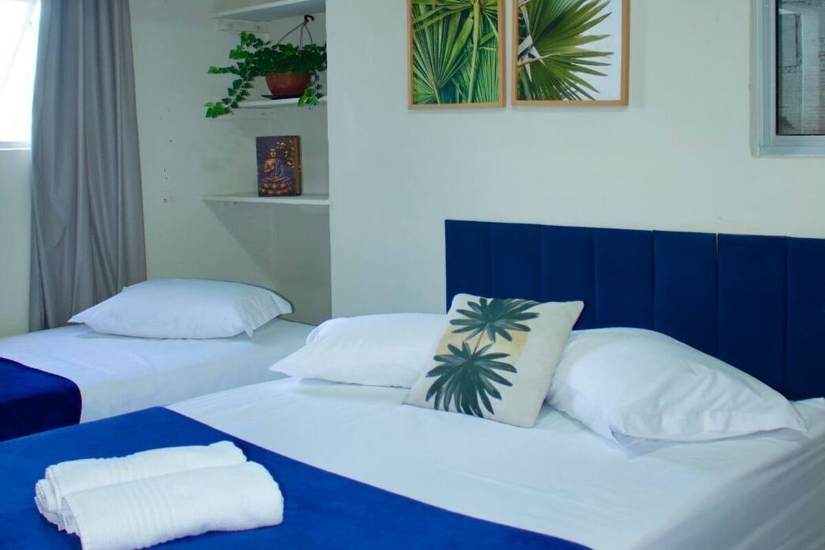 A hotel room with two beds, a blue headboard, a potted plant, and a wall-mounted air conditioner.