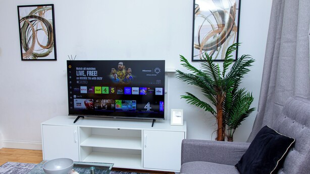 Una sala de estar con una televisión de pantalla plana sobre un soporte blanco, un sofá gris, una mesa de centro de vidrio con un cuenco y un libro, y una planta en maceta.