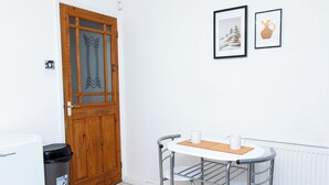 Una habitación pequeña con una puerta de madera, un radiador blanco, una mesita con dos sillas y dos cuadros en la pared.