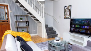 Una sala de estar moderna con un sofá, sillones, una mesa de centro de vidrio, un televisor, una escalera y una estantería.