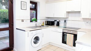 Cocina compacta con armarios blancos, lavadora integrada, microondas y una pequeña ventana con una planta.