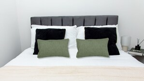 Una cama con cabecera tapizada de gris, almohadas blancas y negras, y una almohada verde.