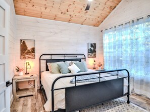 Un dormitorio con una cama, dos mesitas de noche, un tocador, techo de madera y una ventana con cortinas.