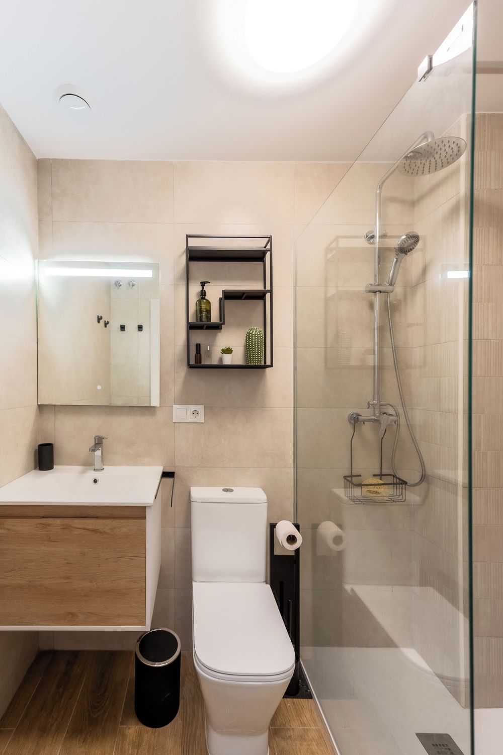 Un baño moderno con ducha envuelta en vidrio, inodoro blanco, tocador de madera y un espejo sobre el lavamanos.