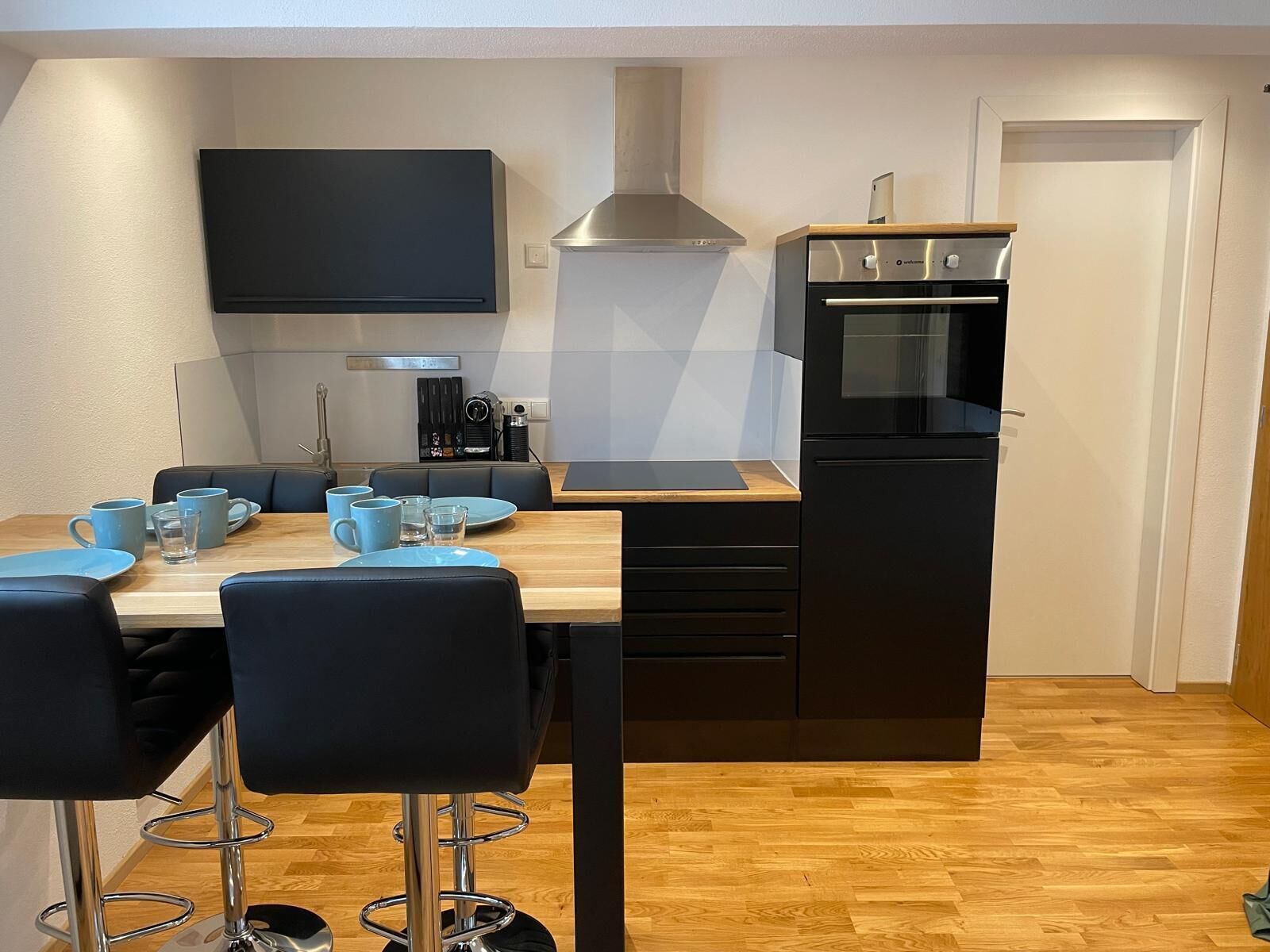 A modern kitchen with a wooden countertop, black cabinets, and a built-in oven. There is a dining area with a table and chairs, and a flat-screen TV mounted on the wall.