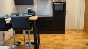 A compact kitchen with a built-in oven, a microwave, and a stove. There are two black bar stools and a wooden dining table with blue cups.