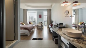 Una habitación de hotel moderna con una cama grande, una zona de estar, un baño con un espejo amplio y un lavamanos, y vistas a la ciudad.