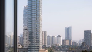 Un edificio moderno de gran altura con fachada de vidrio, rodeado de otros edificios altos, visto desde un balcón con cercado de malla y una silla.
