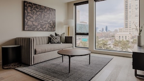 A modern living room with a sofa, a coffee table, a large window with a city view, and a decorative wall piece. - Sosuite at The Onyx - Museum District (Philadelphia)