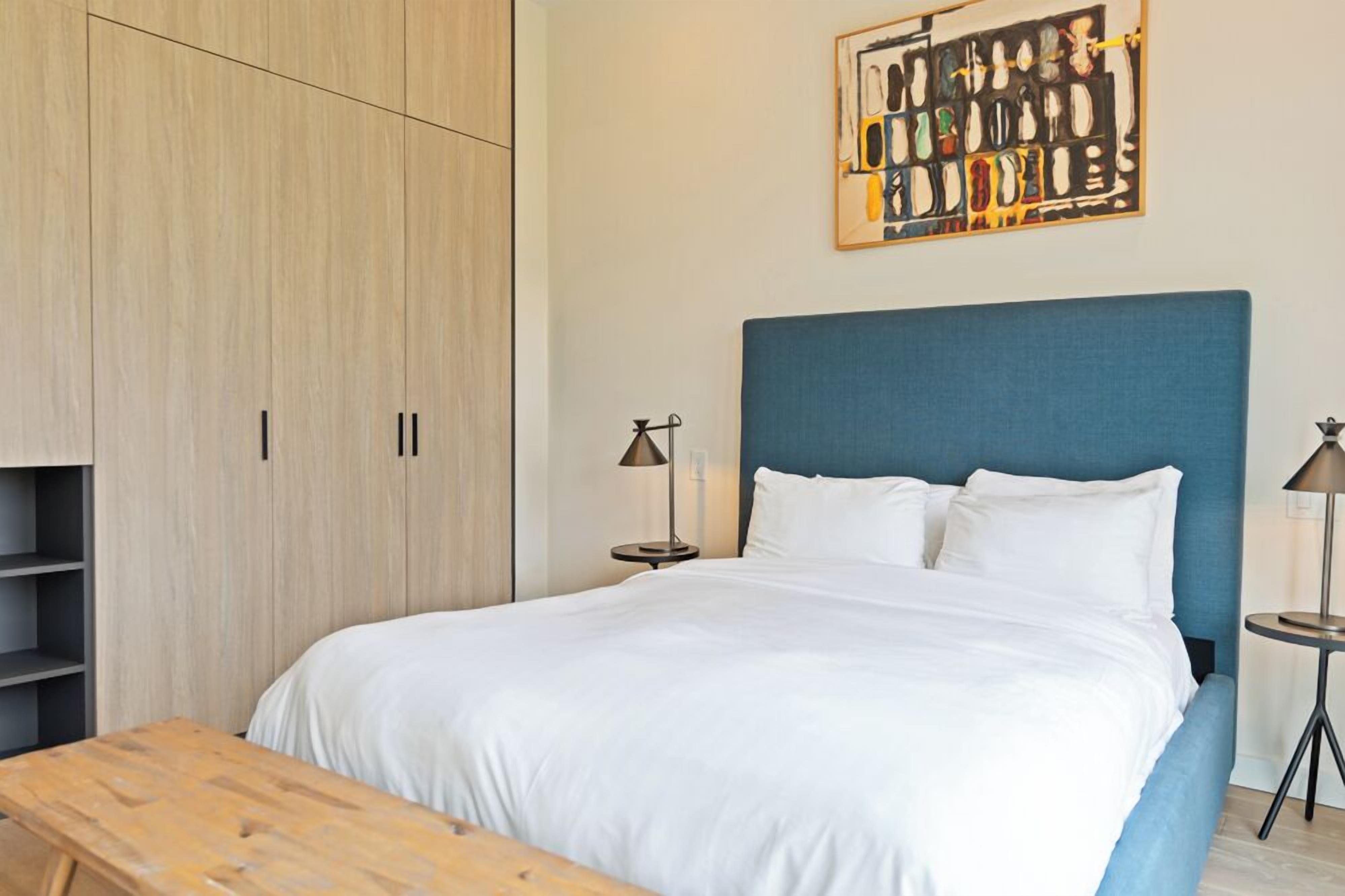 A bedroom with a large bed, a wooden bench, a wardrobe, and a wall-mounted artwork.