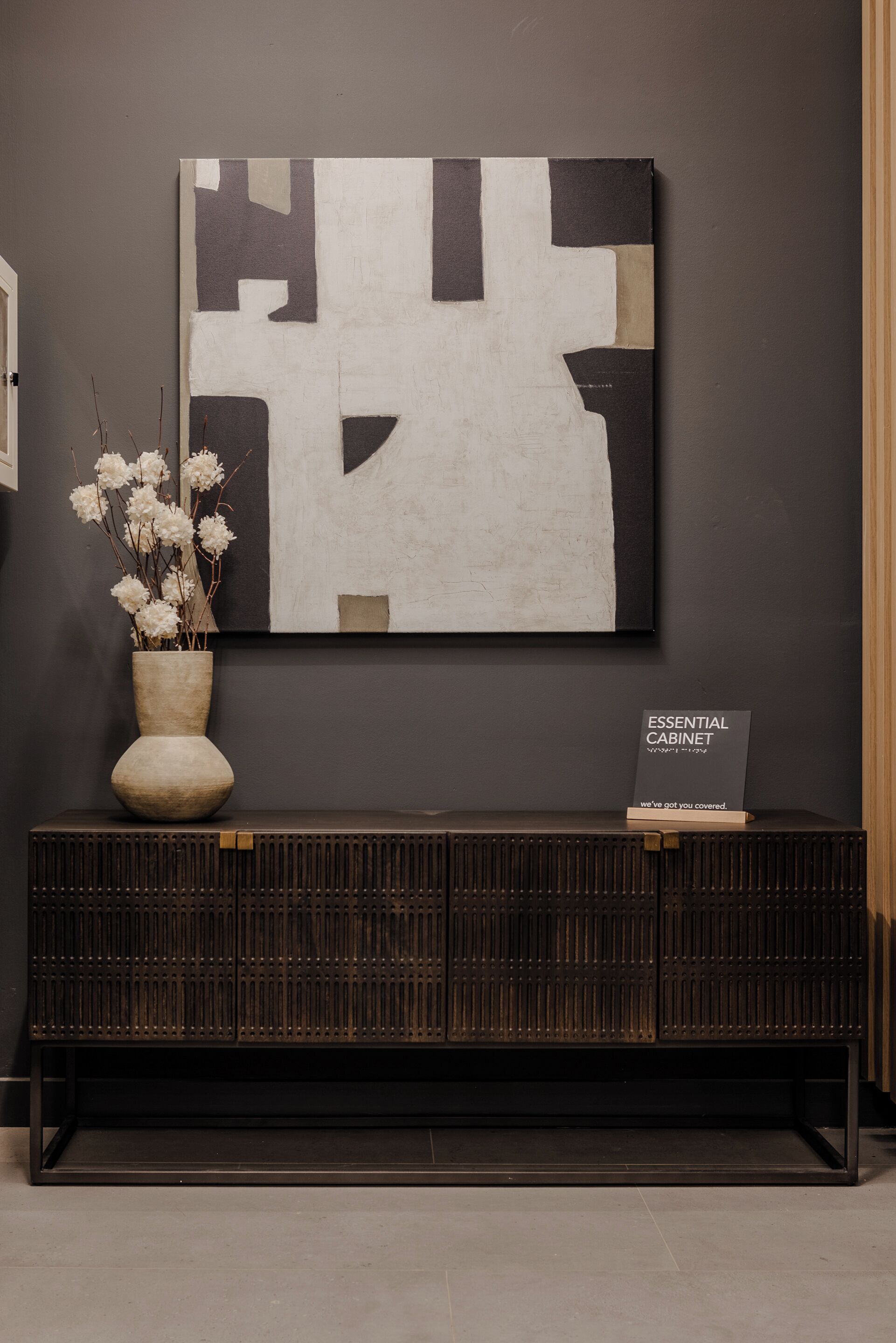 A modern interior with a dark wall, a geometric abstract painting, a wooden console table with a vase of flowers, and a book titled "ESSENTIAL CABINET".
