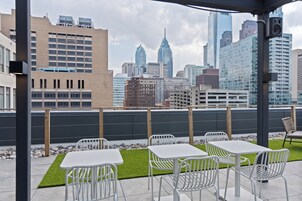 Un espacio al aire libre con mesas y sillas, vista a un horizonte urbano y un edificio moderno con un diseño arquitectónico único.