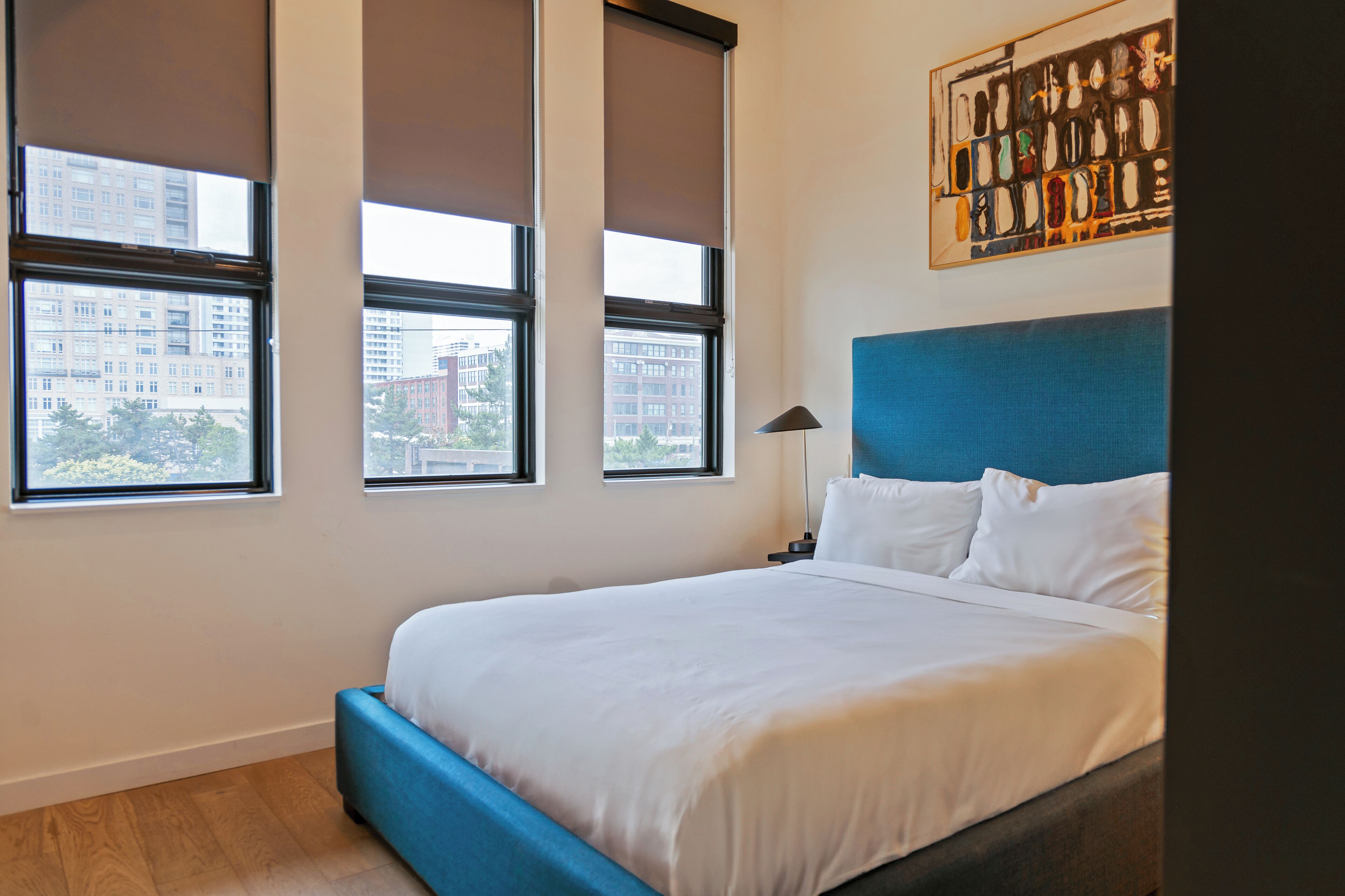 A modern hotel room with a large bed, a blue headboard, and three windows offering a city view.