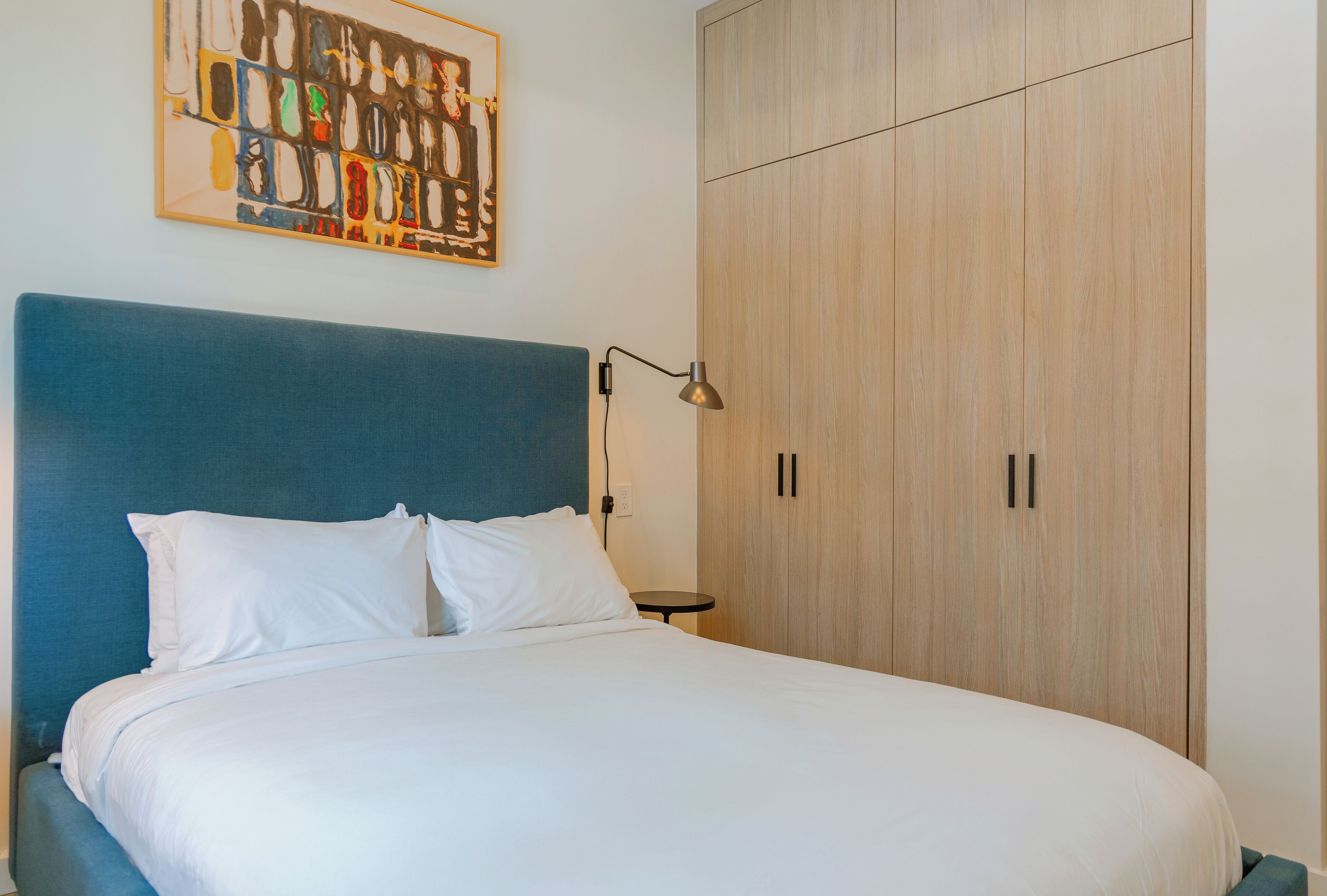 A neatly made bed with white linens, a blue headboard, and a wall-mounted abstract painting.