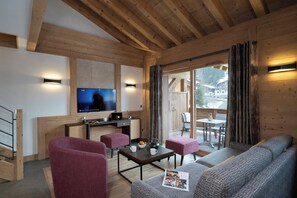 Interior acogedor de una cabaña con techo de madera, un televisor montado en la pared, zona de comedor con mesa y sillas, y un sofá con una revista.