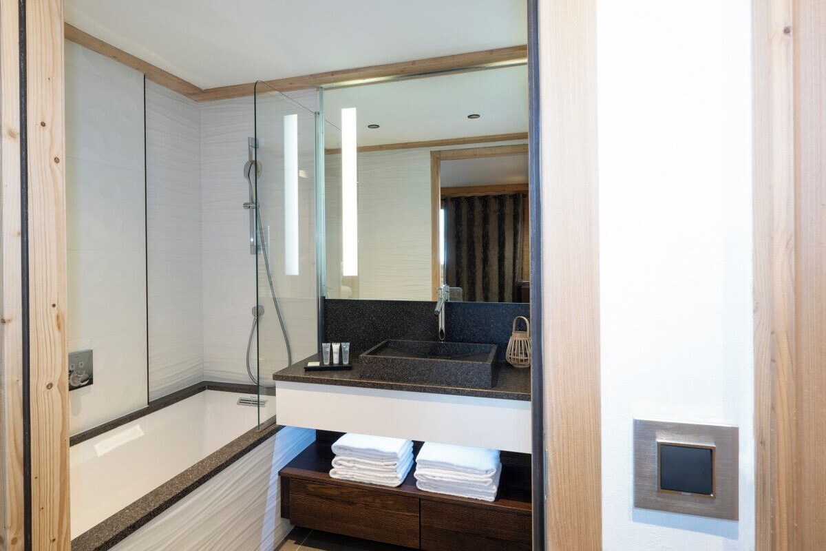 A modern bathroom with a glass-enclosed shower, a sink with a dark countertop, and a mirror above it. There are towels stacked on a shelf below the sink.