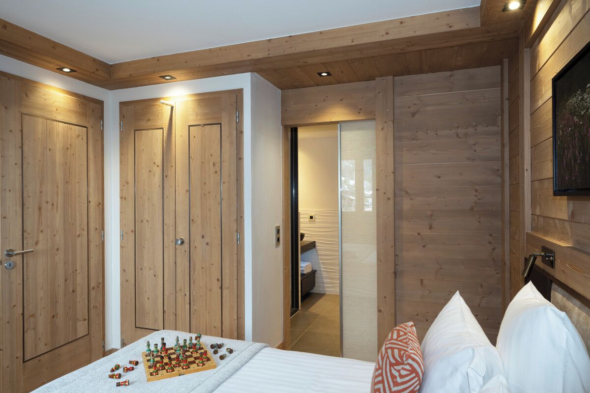 Une chambre avec des murs lambrés en bois, un lit agrémenté d’oreillers blancs et à motifs, et un jeu d’échecs posé sur le lit.