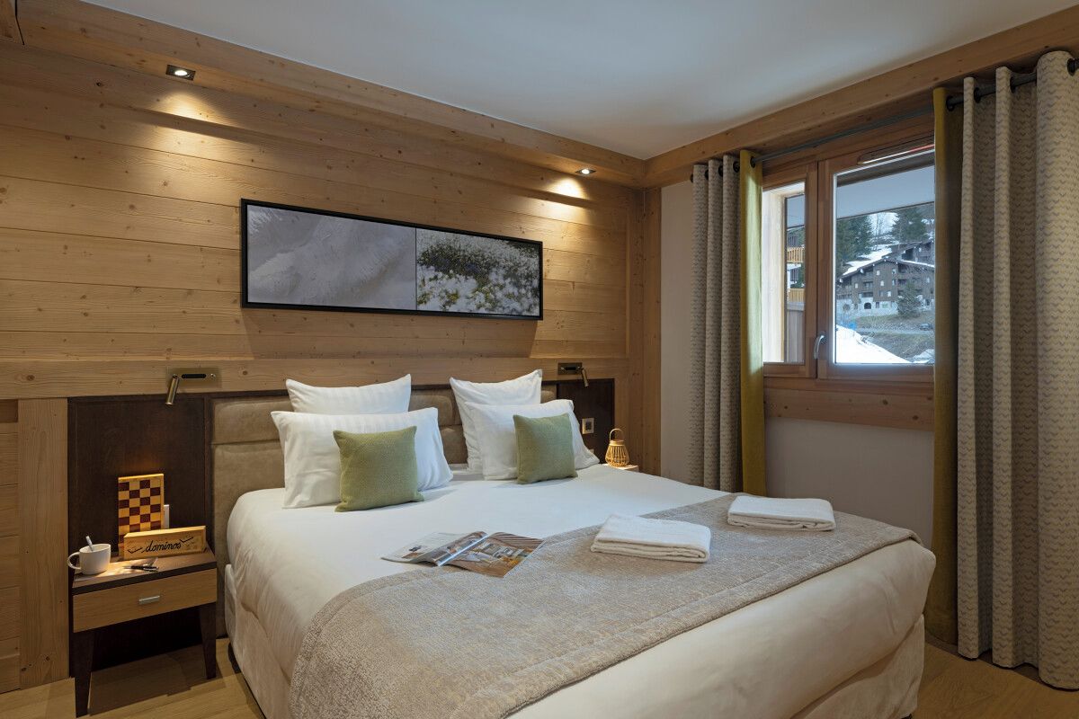 Ein Hotelzimmer mit einem hölzernen Kopfteil, einem großen Bett mit weißen und grünen Kissen, einem Nachttisch mit Lampe und einem mit Vorhängen drapierten Fenster.