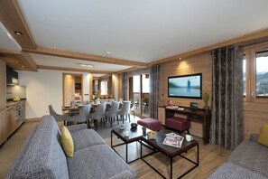 A spacious living area with a sofa, coffee table, and a TV. There is a dining area with chairs and a table, and a kitchen in the background. - MGM Hôtels et Résidences - Residence Anitea (Les Avanchers-Valmorel)