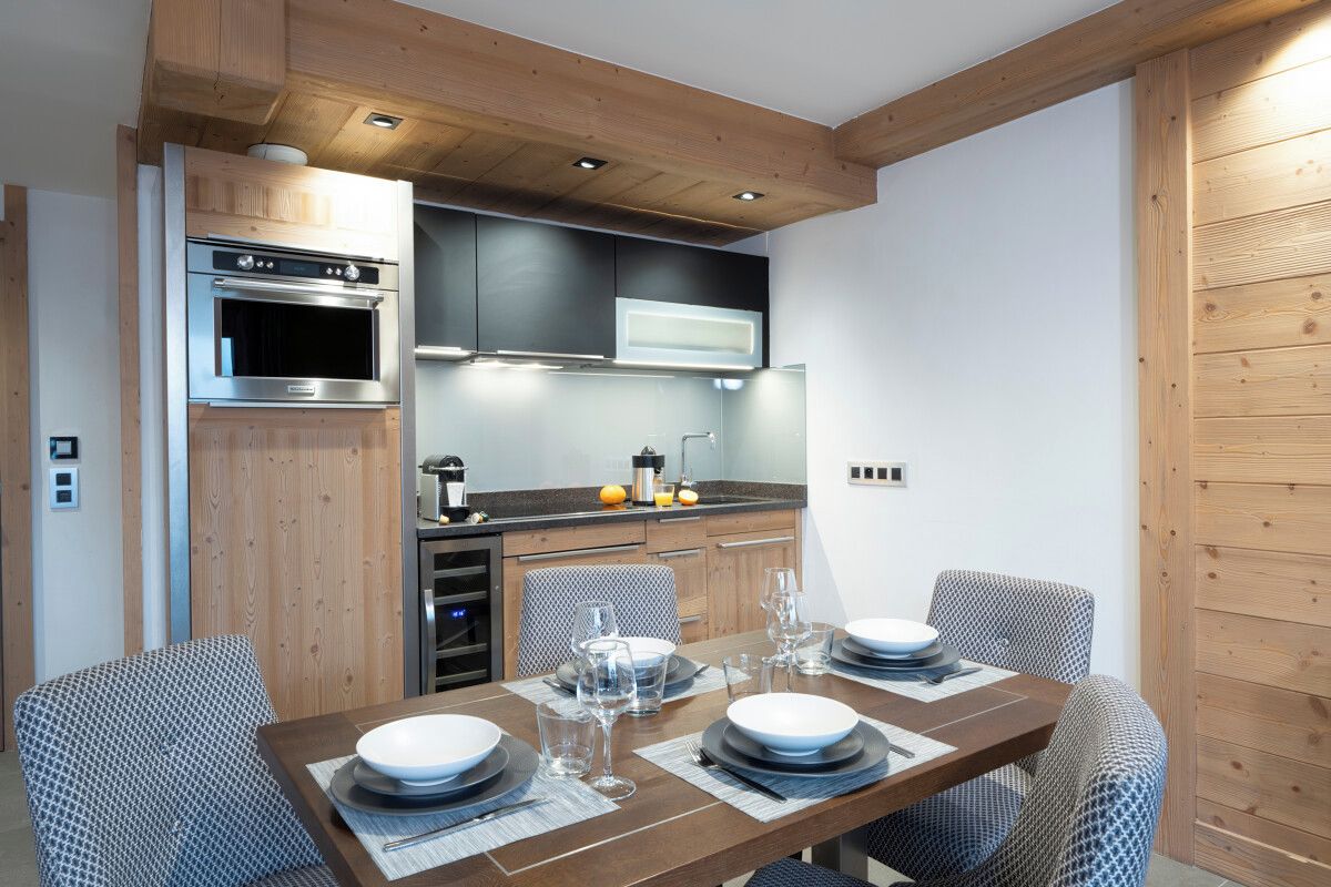 A compact kitchen with built-in appliances, a dining area with a wooden table and patterned chairs, and a wooden ceiling.