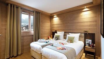 Una habitación con paneles de madera, dos camas, una ventana con cortinas y una mesita de noche con un libro y un peluche.