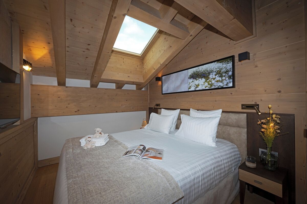 Un dormitorio acogedor con tragaluz, una cama con ropa de cama blanca, una mesita de noche con un jarrón y un cuadro en la pared.