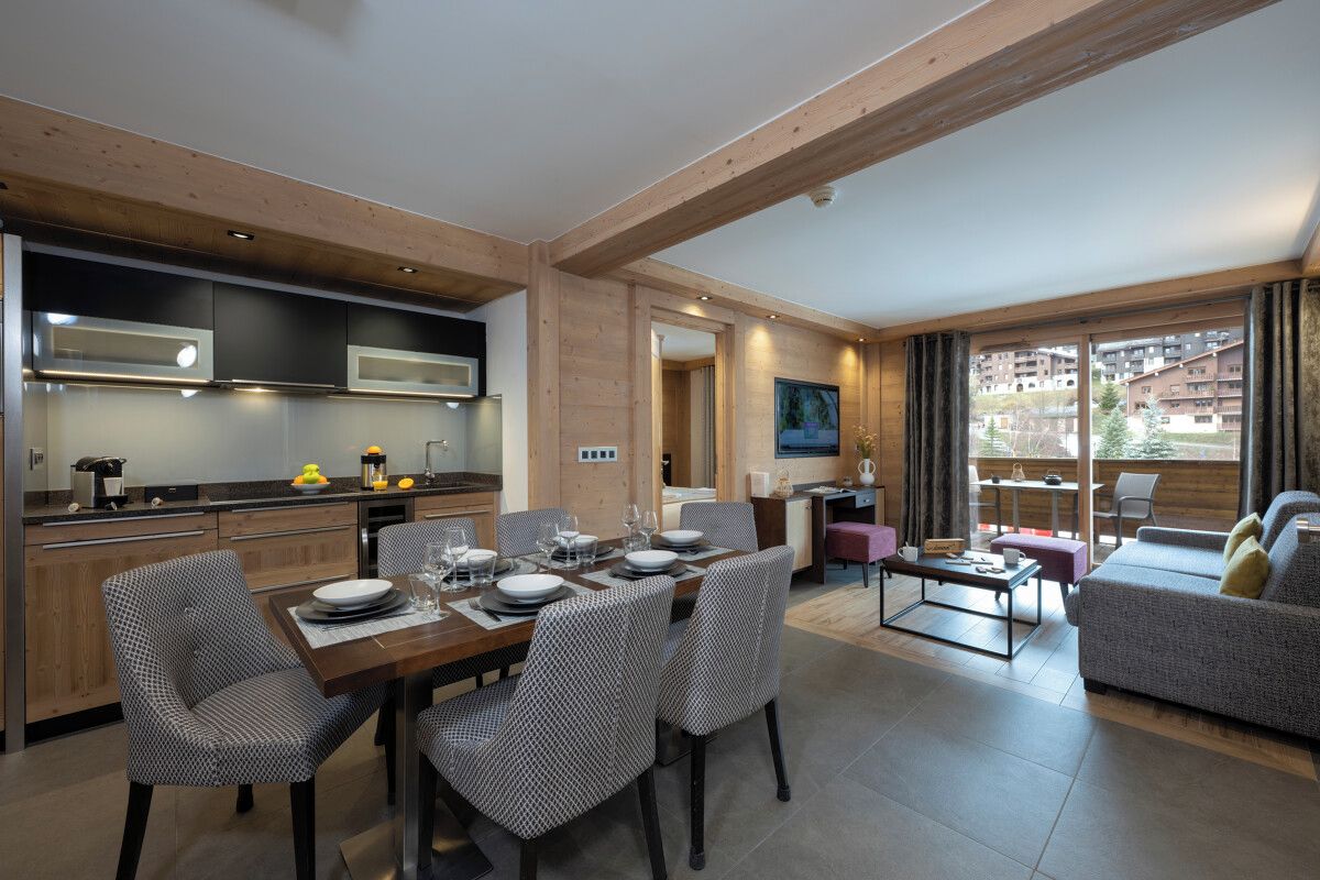 A modern kitchen with wooden beams, a dining area with a table set for four, and a living area with a sofa and a balcony with a view of buildings.