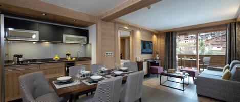 Une cuisine moderne avec des poutres en bois, un coin repas avec une table dressée pour quatre personnes, et un salon avec un canapé et un balcon offrant une vue sur les bâtiments.