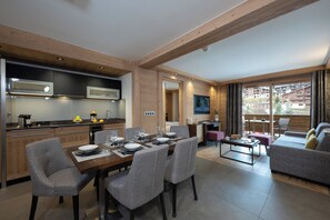 A modern kitchen with wooden beams, a dining area with a table set for four, and a living area with a sofa and a balcony with a view of buildings. - MGM Hôtels et Résidences - Residence Anitea (Les Avanchers-Valmorel)