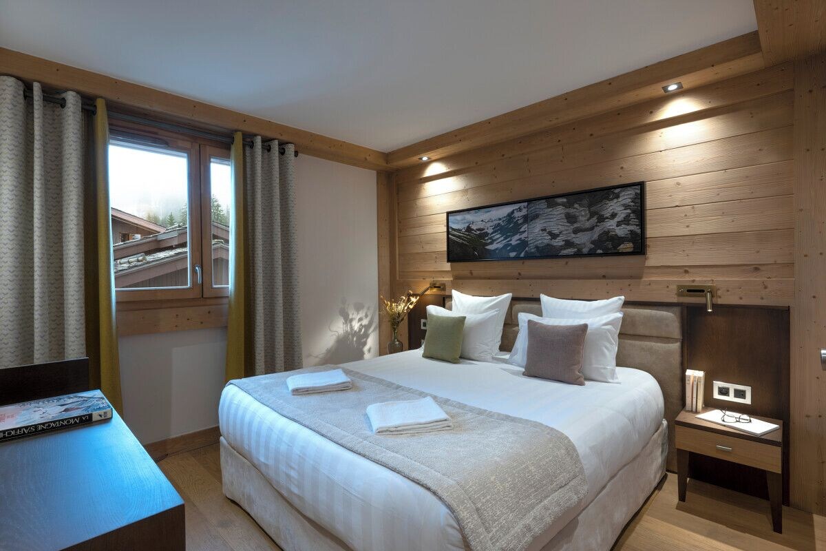 A neatly arranged bedroom with a large bed, a bedside table, a nightstand, a window with curtains, and a framed picture on the wall.