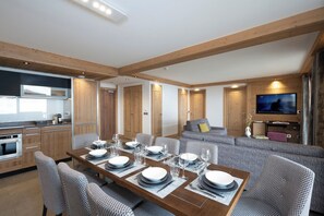 Un salon moderne comprenant un coin repas, des poutres en bois, un téléviseur et une cuisine équipée d’appareils électroménagers intégrés.
