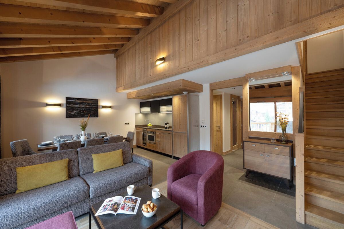Un salon moderne avec un canapé, des fauteuils et une table basse. On y trouve également un coin cuisine avec des meubles en bois et un coin repas avec des tables et des chaises.