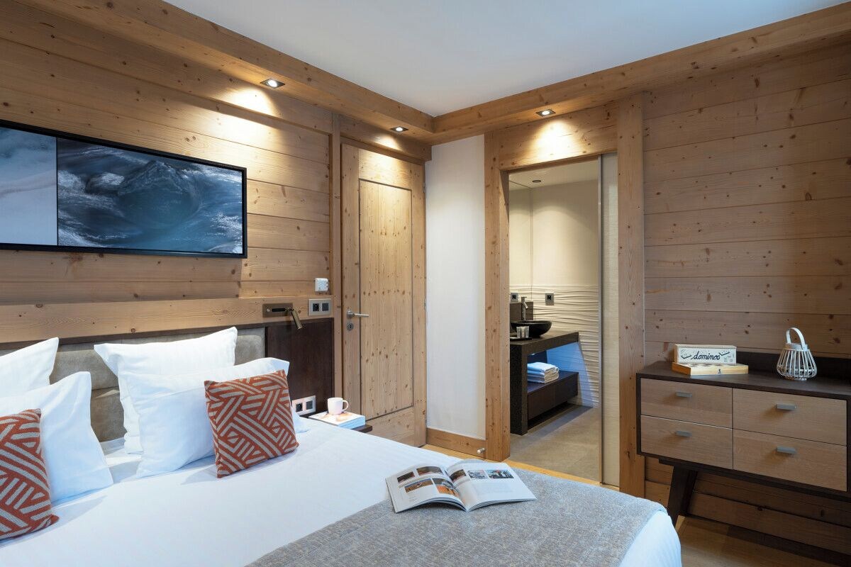 Une chambre d’hôtel avec un intérieur en bois, un lit agrémenté d’oreillers blancs et à motifs, une table de chevet avec un livre, et une vue sur la salle de bain.