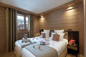 A cozy room with wooden walls, two beds with pillows, a nightstand with a lamp, and a window with curtains. - MGM Hôtels et Résidences - Residence Anitea (Les Avanchers-Valmorel)