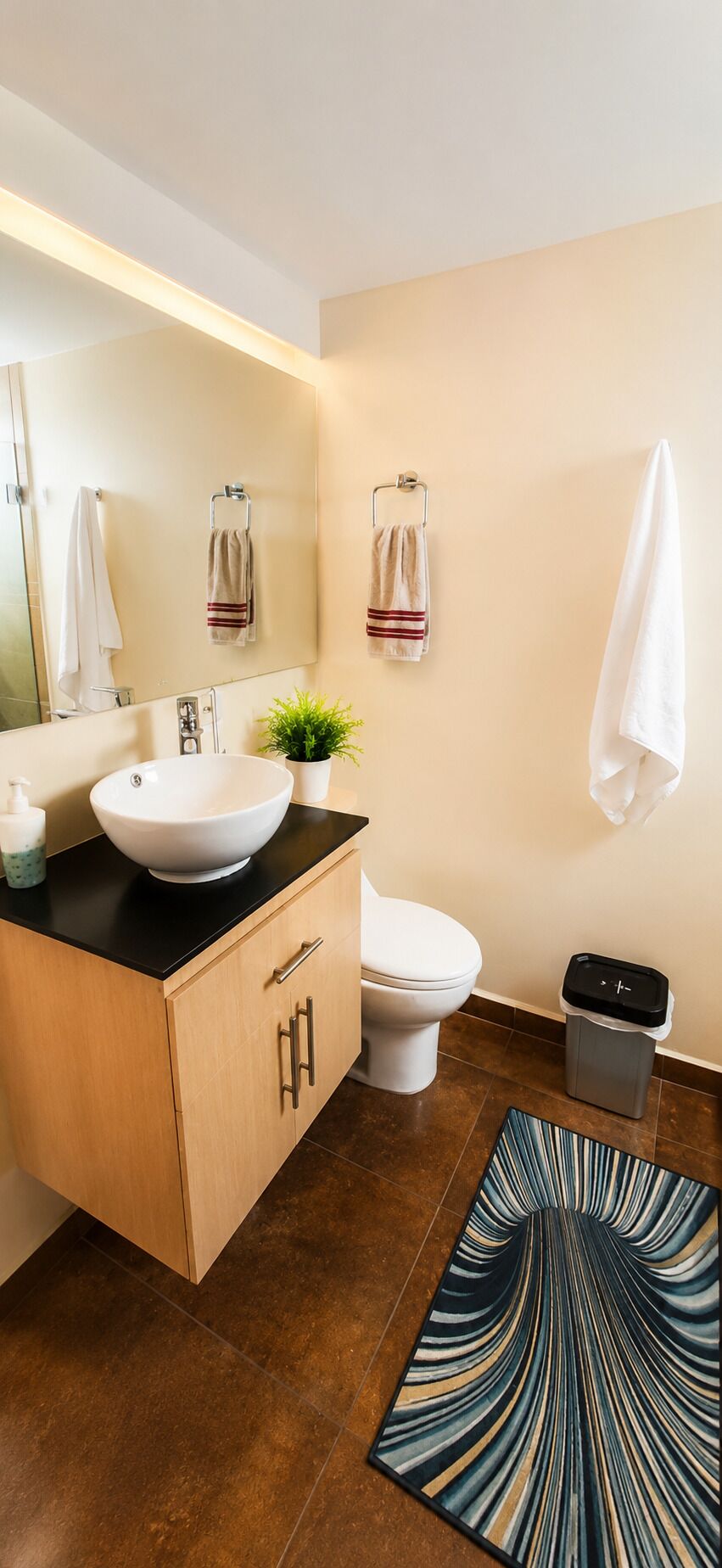 A modern bathroom with a large mirror, a white sink, a toilet, and a trash bin.