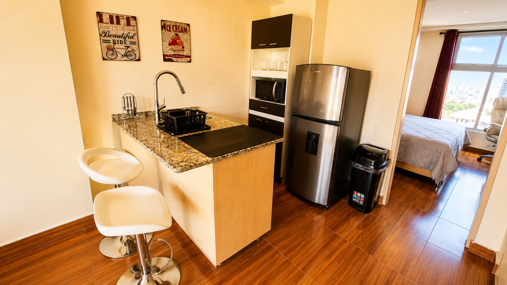 A compact kitchen with a built-in refrigerator, a microwave, and a sink. There are two white bar stools and a small island with a granite countertop. The room has wooden flooring and a large window with a view.