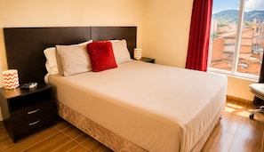 A neatly made bed with a red accent pillow, a bedside lamp, and a nightstand with a remote control. - Comfortable  apartment Great location (Medellín)