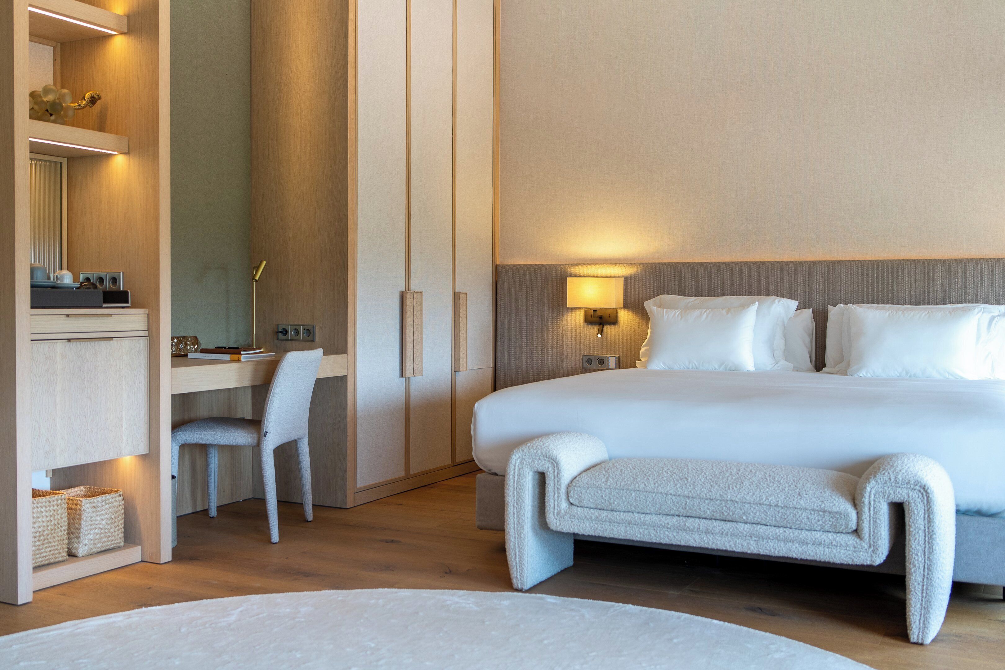 A modern hotel room with a large bed, a desk with a chair, a bedside table, and a wall-mounted lamp.