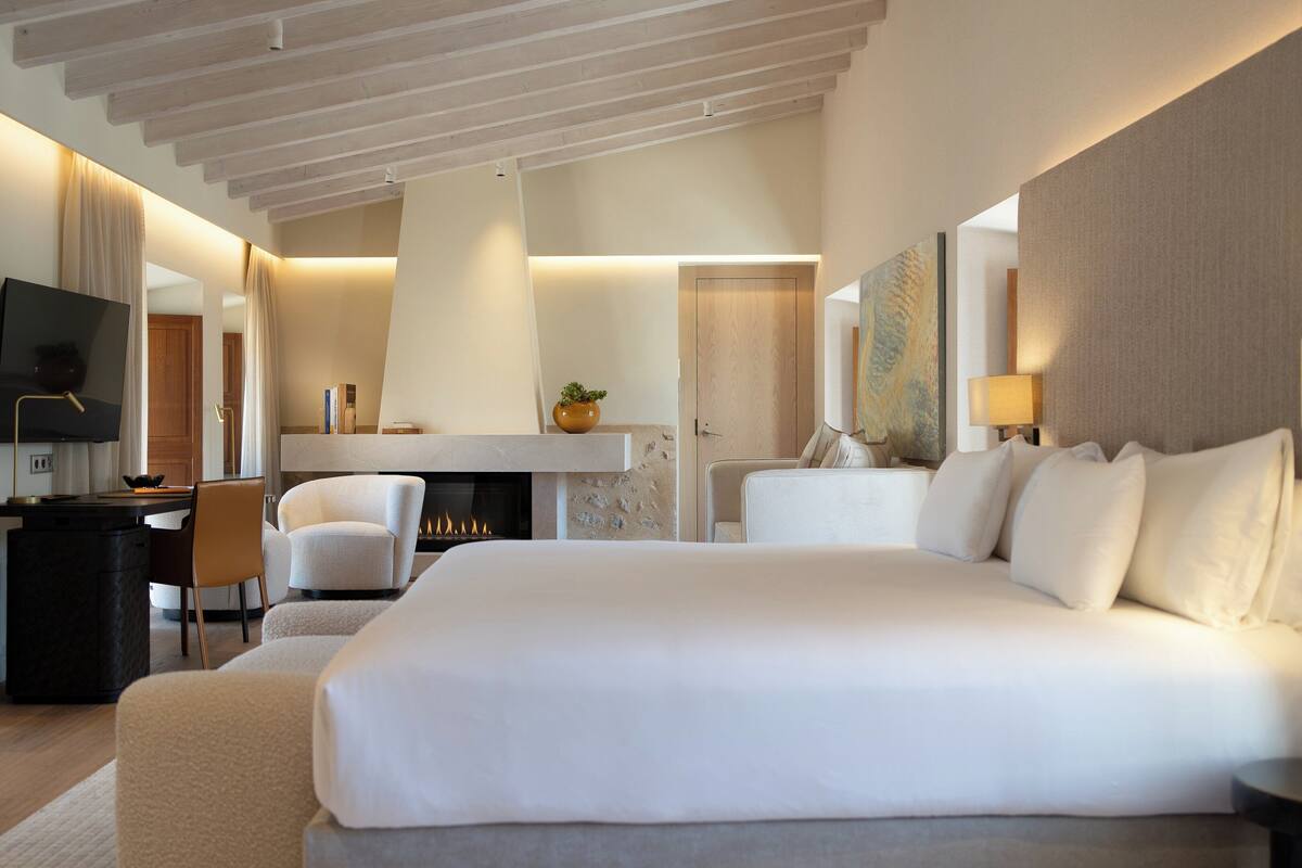 A modern hotel room with a large bed, a desk, a chair, a TV, a fireplace, and a painting on the wall.
