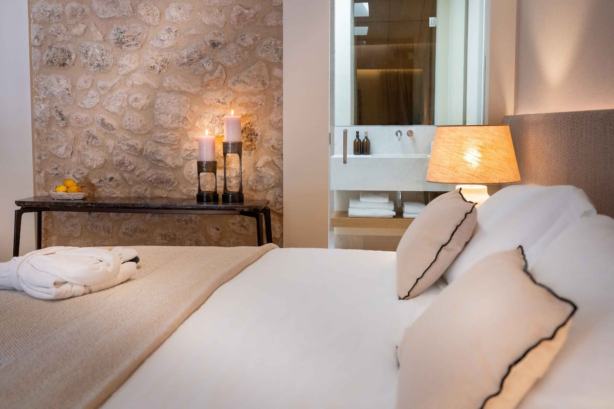 A neatly made bed with pillows, a bedside table with candles, a mirror, and a lamp.