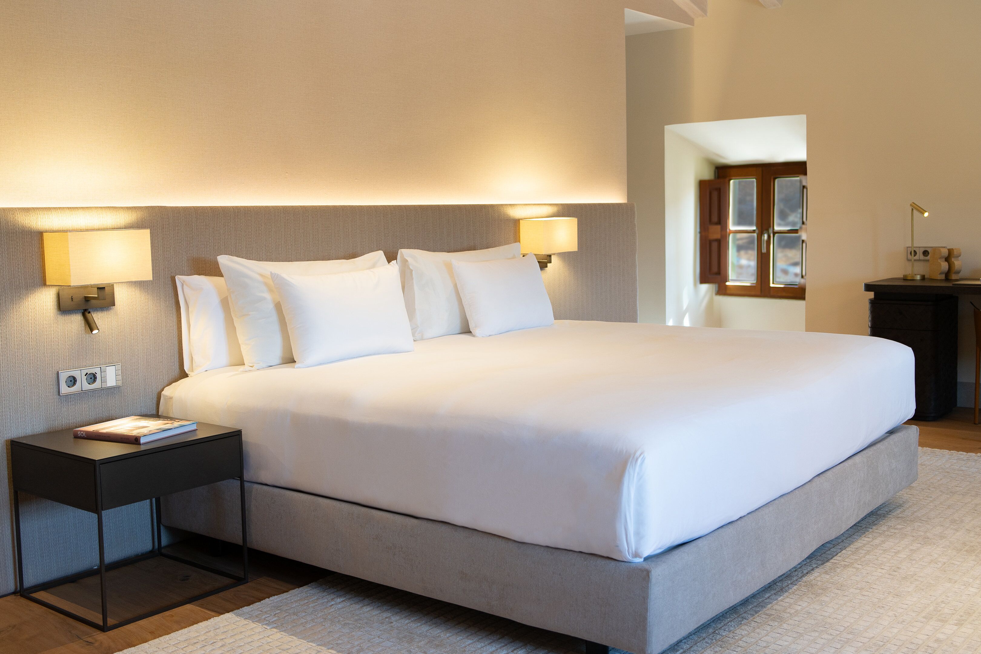 A neatly made bed with white linens, two bedside lamps, and a small bedside table with a book.